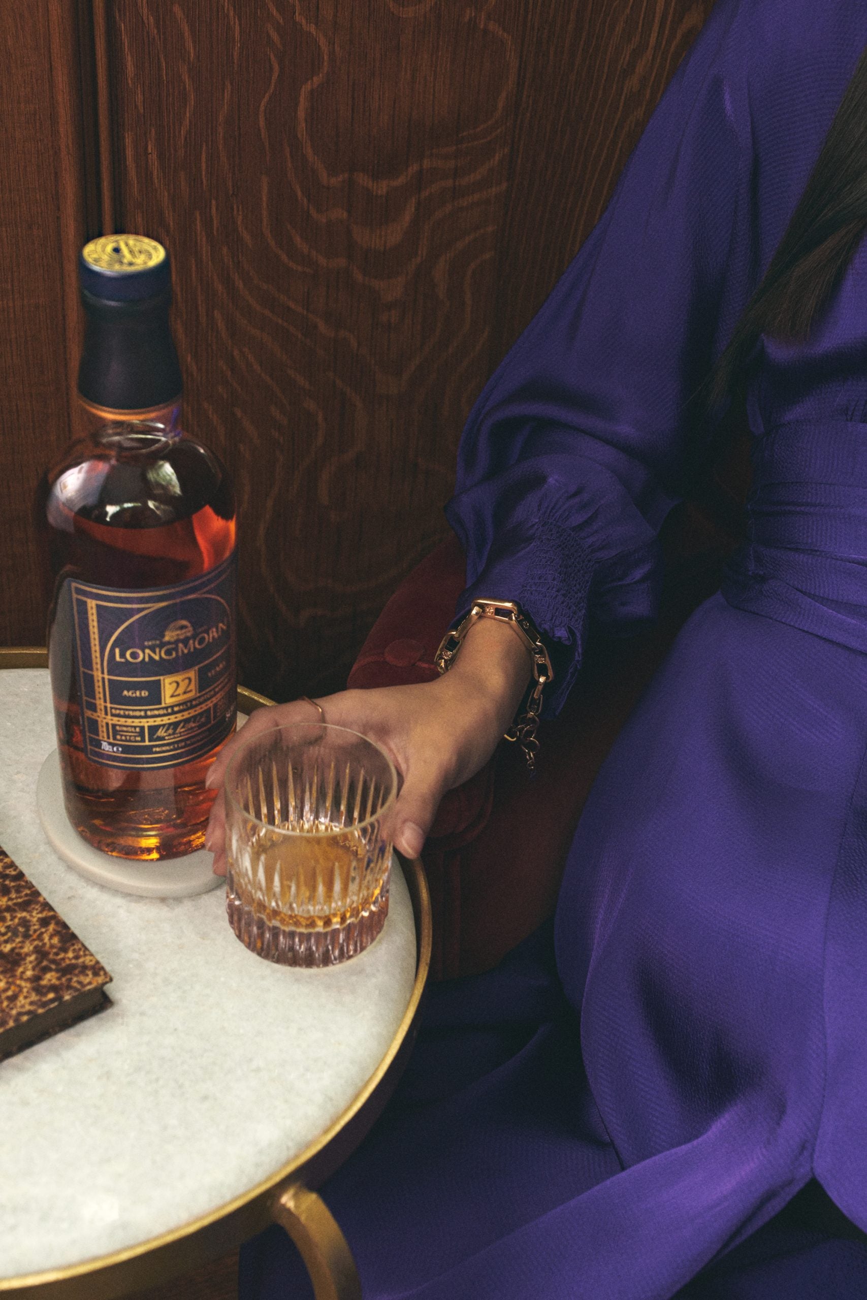 A woman in purple holding a glass of whisky next to a bottle of 22 Year Old Longmorn Whisky