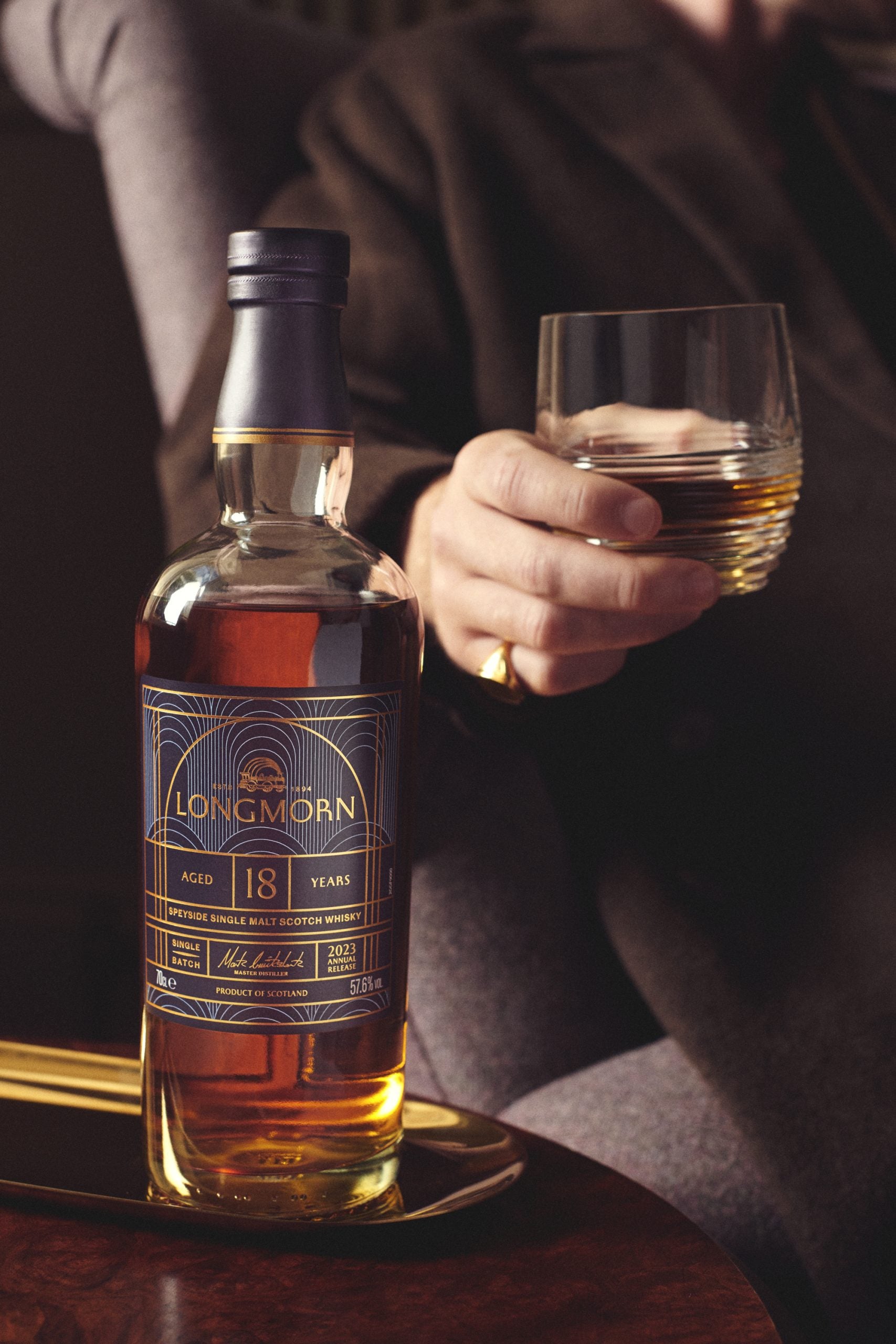 A hand holding a glass of whisky with a bottle of 18 Year Old Longmorn Whisky